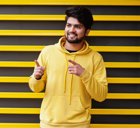 urban-young-hipster-indian-man-fashionable-yellow-sweatshirt-cool-south-asian-guy-wear-hoodie-against-striped-background-showing-fingers-free-space 1